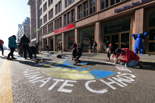 Eine Gruppe von Menschen sitzt vor einem Gebäude mit Fenstern und Namensschildern auf dem Boden, umgeben von Flaschen und anderen Gegenständen, während sie an einer Klimawandel-Demonstration in Berlin teilnehmen, die von Bäumen und einem klaren blauen Himmel umgeben ist.