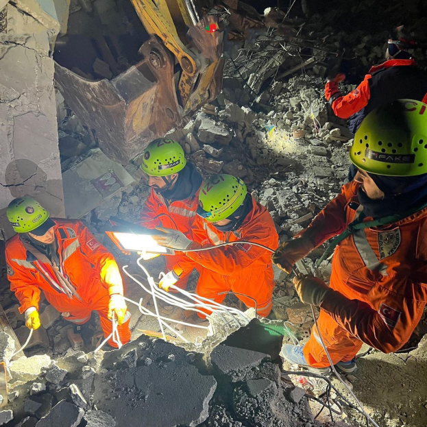 Eine Gruppe von Menschen in orangefarbigen Uniformen und grünen Helmen bei der Arbeit an Schutt von einem eingestürzten Gebäude, mit Werkzeugen und Materialien, im Hintergrund ein Kran.