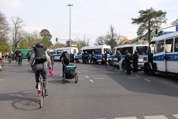 Eine Gruppe von Menschen, die auf Fahrrädern eine Straße entlangfahren, mit Polizeiwagen, anderen Fahrzeugen, Bäumen, Laternen, Verkehrszeichen, Schildern, Gebäuden und einem bewölktem Himmel.