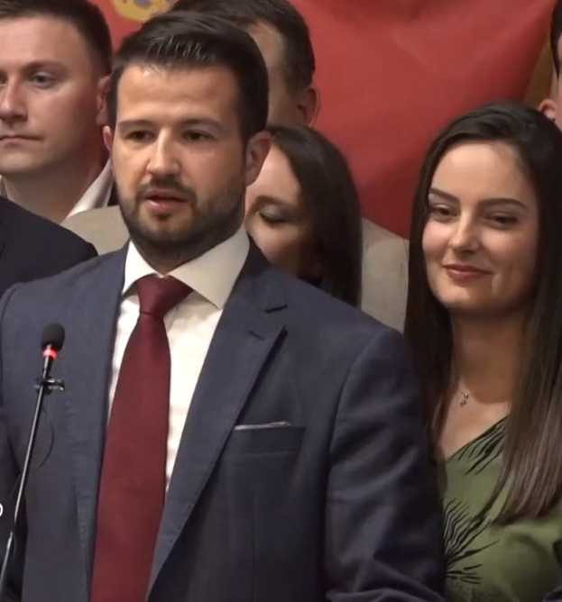 Ein Mann im Anzug spricht in ein Mikrofon auf einem Podium, mit einem Banner dahinter und einer Menge im Hintergrund, wahrscheinlich zu einem Regierungsverbot von Social Media.