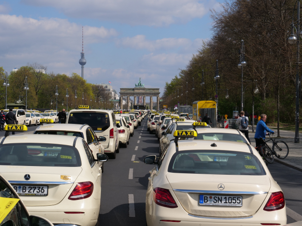 Eine lange Reihe von Taxis, die an der Seite einer belebten Straße in Berlin, Deutschland, geparkt sind, mit Fahrzeugen, Fahrradfahrern und Füügängern, flankiert von Laternenmästen, Bäumen und Gebäuden, einschließlich eines Bogens und eines Turms.