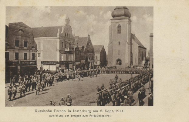 Ein Schwarz-Weiß-Foto einer Parade in Insterburg am 5. September 1914 mit vielen Menschen, Gebäuden im Hintergrund und Text am unteren Bildrand.