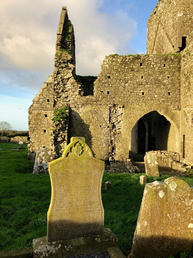 Ruinen einer alten Kirche in Irland mit einem Friedhof im Vordergrund und einer Burg im Hintergrund bei bewölktem Himmel, mit einem Wasserzeichen.