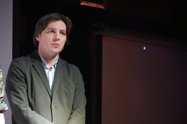 Zwei Personen nebeneinander auf einer Bühne, eine in einem Anzug und die andere neben ihnen, beide mit ernsten Gesichtern.