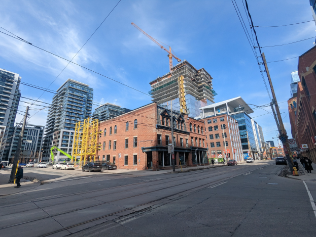 Eine belebte Stadtstraße mit Fahrzeugen, Fußgängern, Strommasten, Schildern und Gebäuden, mit einer Baustelle in Toronto im Hintergrund unter einem bewölkten Himmel.