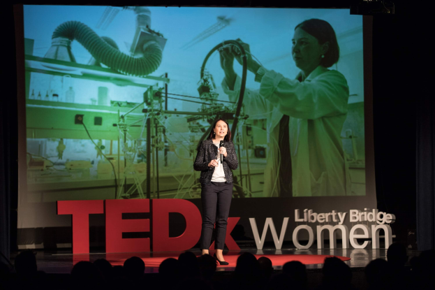 Frau auf einer Bühne mit Mikrofon, hinter ihr ein Display mit Bild und Text zum TEDxWomen-Event.