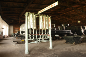 Ein Bahnsteig mit einem Zug auf den Gleisen, einem Rollstuhl auf dem Boden, einer Signalbox in der Mitte, einem Pfahl mit einer Tafel daran, einer Wand links und einer Decke darüber.