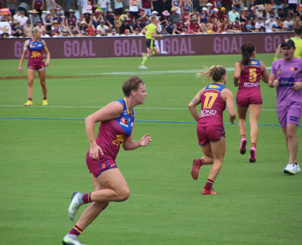 Frauen spielen Australian-Rules-Football auf einem Rasenfeld mit Zuschauern im Hintergrund.