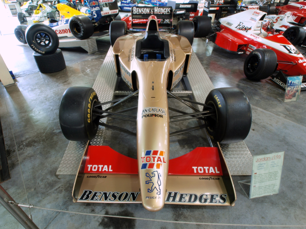 Rennwagen in einem Museum hinter einem Metallzaun, mit einer Texttafel und Menschen im Hintergrund.