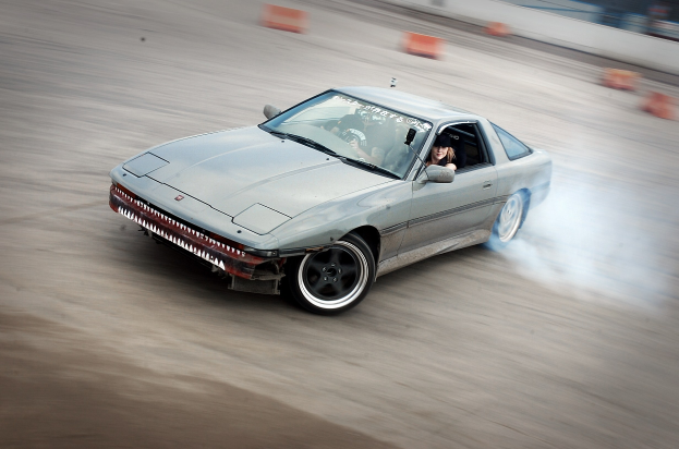 Eine Person in einem Helm fährt ein Auto, das Rauch ausstößt, auf einer von orangefarbenen Objekten umgebenen Strecke, mit einem unscharfen Hintergrund, der hohe Geschwindigkeit andeutet.