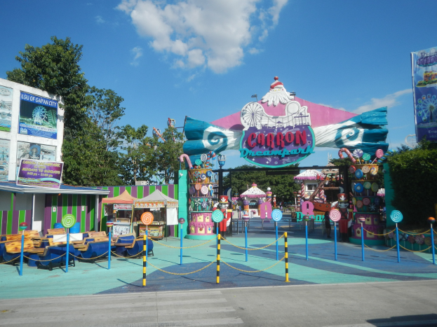 Kirmesfahrgeschäft am Parkeingang mit Straße, Umzäunung, Buden, Schildern, Bäumen und bewölktem Himmel im Hintergrund.
