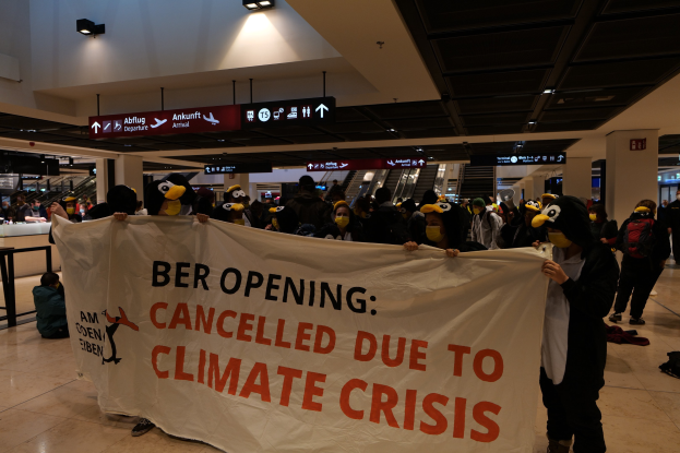 Gruppe von Menschen mit Masken, die ein Banner mit der Aufschrift "Ber-Eröffnung wegen Klimakrise abgesagt" tragen, in einem Flughafen, mit einigen stehenden und anderen sitzend auf dem Boden, Rolltreppen und Schilder im Hintergrund.