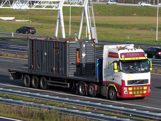 Ein Lastwagen mit einem großen Behälter fährt auf einer Autobahn, mit anderen Fahrzeugen, Polen, Bäumen, Gebäuden und einem klaren blauen Himmel im Hintergrund.
