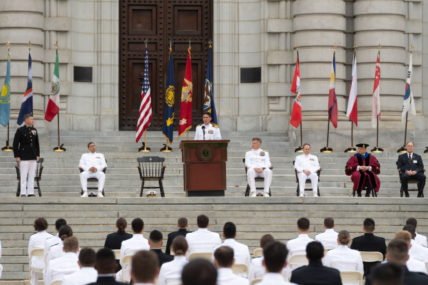 Eine Gruppe von Menschen in weißen Uniformen sitzt auf den Stufen eines Gebäudes, mit einem Podium und Fahnen davor und einem großen Gebäude mit einer Tür im Hintergrund.