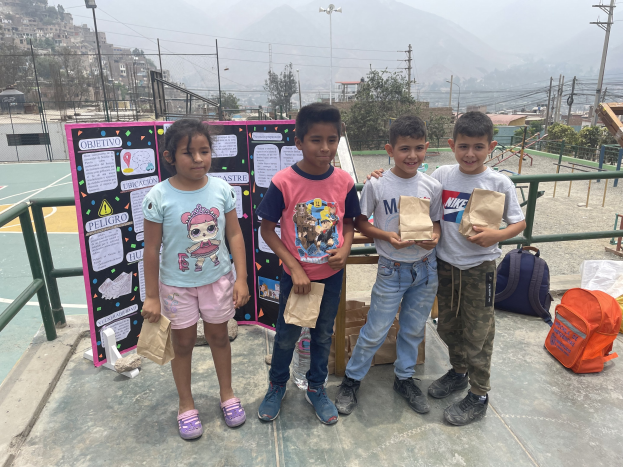 Gruppe von Kindern auf einem Basketballfeld mit Papierbeuteln in den Händen, Gegenstände auf dem Boden rechts und eine Tafel mit Text links, vor einer Außenkulisse mit Zaun, Pfosten, Gebäuden.
