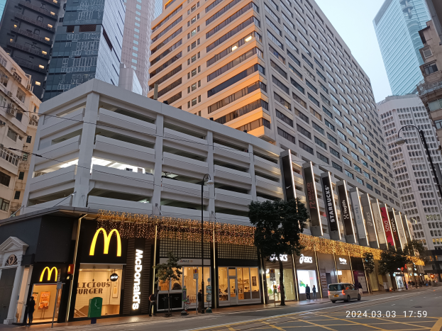 Ein McDonald's-Restaurant auf einer belebten Stadtstraße, flankiert von hohen Gebäuden, Straßenlaternen und -masten, mit Fahrzeugen auf der Straße, Fußgängern auf dem Gehweg, Bäumen und einem klaren blauen Himmel.