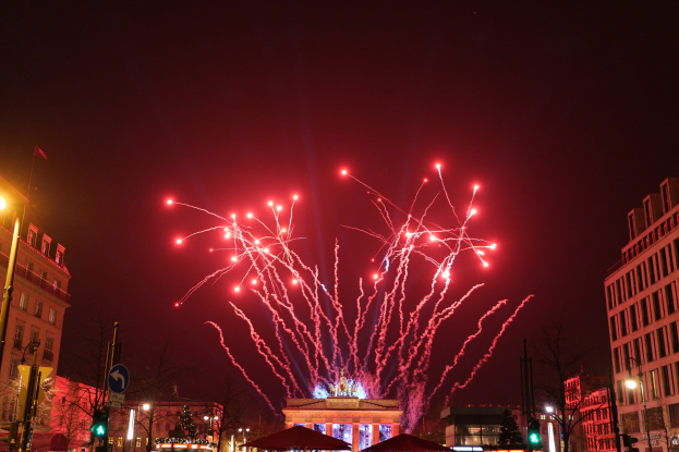 Eine belebte Stadtstraße an einem Silvesterabend in Berlin, mit Gebäuden, Bäumen, Laternen, Verkehrszeichen, Schildern, Zelten, Menschen und einem prächtigen Feuerwerk am Himmel.