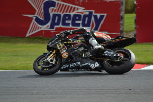 Eine Person in Helm und Schuhwerk fährt ein Motorrad auf einer von Gras umgebenen Rennstrecke, mit einer Tafel im Hintergrund, auf der Text zu sehen ist.