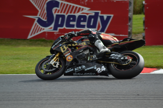 Eine Person in Helm und Schuhwerk fährt ein Motorrad auf einer von Gras umgebenen Rennstrecke, mit einer Tafel im Hintergrund, auf der Text zu sehen ist.