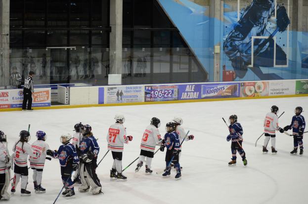 Gruppe junger Menschen, die Eis hockey auf einer Indoor-Eisbahn spielen, mit Helmen und Stöcken und einer Person links daneben und Bannern, Säulen und Glaswänden im Hintergrund.