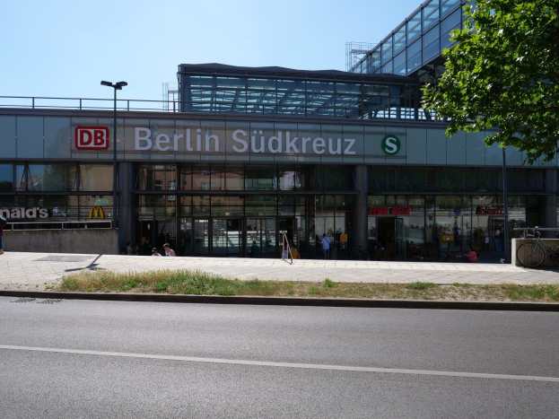 Große Glasfront-Bahnhof Berlin Südkreuz mit sichtbarem Text, umgeben von Straßenmasten, Lichtern, Fahrzeugen, Fütterern, Fahrrädern, Bäumen und einem klaren blauen Himmel.