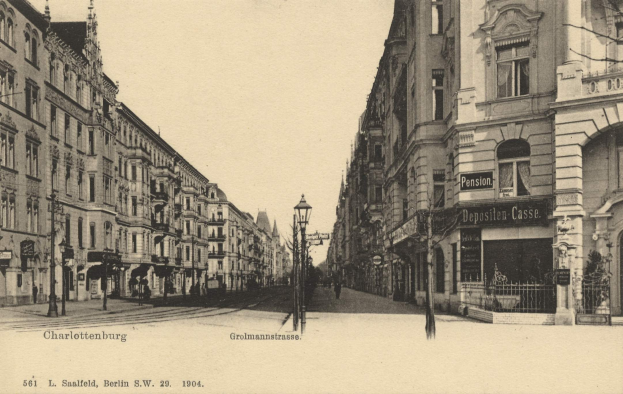 Schwarzes und weißes Foto einer Stadtstraße in Charlottenburg, Deutschland, mit Gebäuden, Straßenlaternen, Lampen, Namensschildern, Fahrzeugen, Fußgängern und Gittern unter einem klaren Himmel.