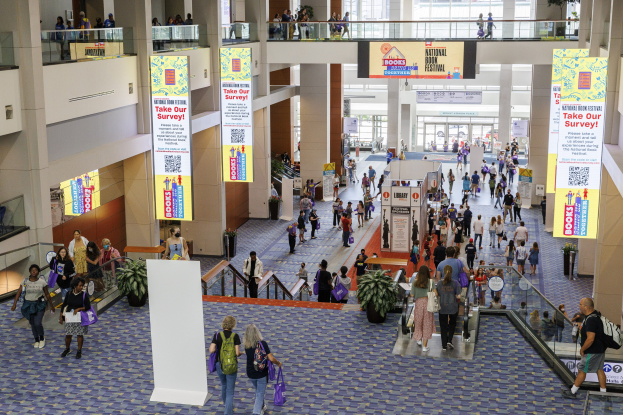 Eine große Gruppe von Menschen, die durch ein großes Gebäude mit Bannern, Topfpflanzen, Rolltreppen, Geländern, Säulen, bedruckten Tafeln, Glastüren und anderen Gegenständen geht, einige tragen Taschen, wahrscheinlich beim Nationalen Buchfestival.