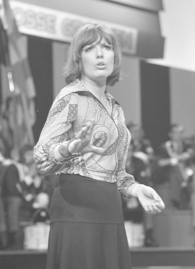 Eine Frau in einem Schwarz-Weiß-Foto steht auf einer Bühne, trägt ein weißes Hemd und einen schwarzen Rock, hat ihre Haare zu einem Dutt gebunden und einen ernsten Ausdruck, umgeben von einer Gruppe von Menschen und einer Wand im Hintergrund.