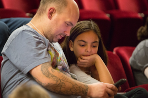 Zwei Menschen, ein Mann und ein junges Mädchen, sitzen in einem Kino, beide konzentrieren sich auf ein Handy; der Mann hält es, während das Mädchen genau hinschaut, mit anderen unscharfen Zuschauern im Hintergrund.