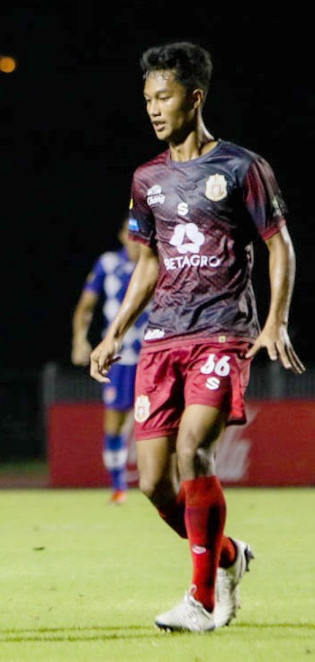 Ein Mann in einem rotbraunen T-Shirt und roten Shorts spielt Fußball auf einem Feld, im Hintergrund ist eine weitere Person und eine Tafel mit Text zu sehen.