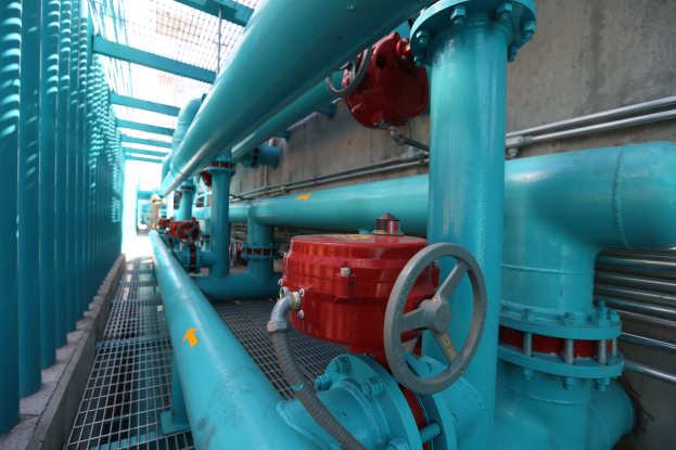 Eine große Gruppe von blauen Rohren und Ventilen in einem Gebäude, mit einem roten Ventil in der Mitte, Metallstangen links und einer Wand im Hintergrund, die wahrscheinlich den Bau einer Gasleitung darstellt.