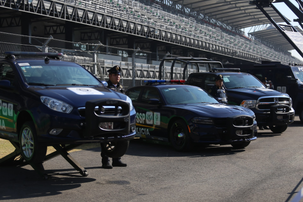Eine Gruppe von Polizeiwagen vor einer Rennstrecke geparkt, mit uniformierten Beamten auf der Straße stehend, im Hintergrund ein Stadion und eine Absperrung.