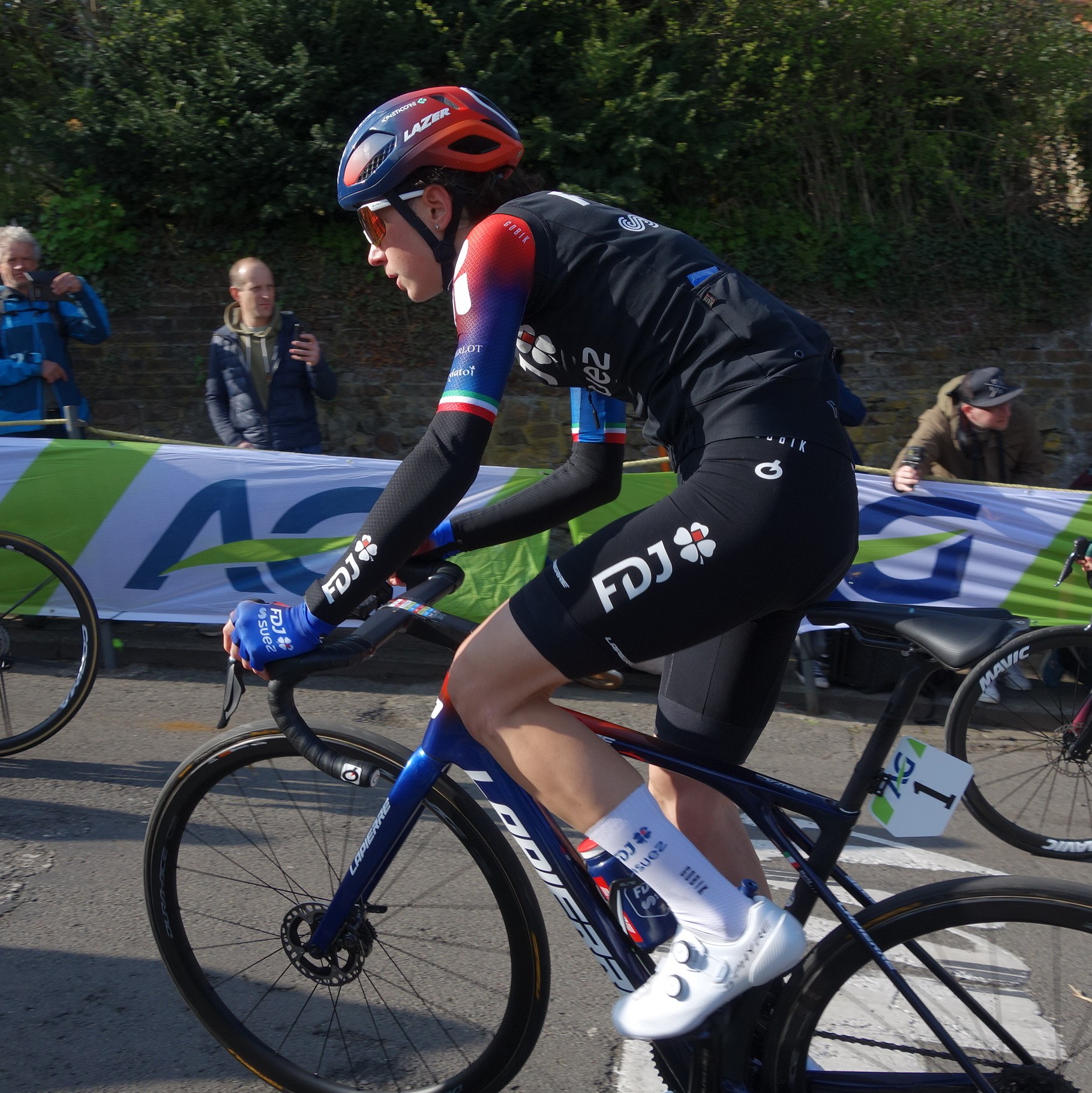 Eine professionelle Radfahrerin, Katie Archibald, trägt einen Helm, eine Schutzbrille, Handschuhe und Schuhe und fährt ein Fahrrad auf einer Straße neben einer Menschenmenge, mit einem Banner, Bäumen, einer Wand und einem Gebäude im Hintergrund.