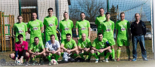 Eine Gruppe von Männern in grünen T-Shirts steht auf einem Fußballfeld mit einem Ball davor, einige in einer Hockposition, in der Nähe eines Torpfostens mit Gebäuden und Bäumen im Hintergrund.