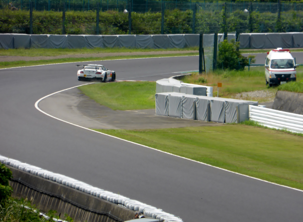 Ein Rennwagen fährt auf einer Rennstrecke mit einem Polizeiwagen im Hintergrund, umgeben von Gras, Pflanzen, Bäumen und einer Umrandung, mit einem Fahrzeug auf der rechten Seite und Text unten.