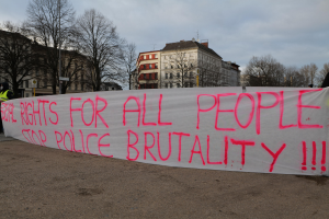 Eine Gruppe von Menschen, die auf dem Boden stehen und ein Banner halten, auf dem "Rechte für alle Menschen Stoppt Polizeigewalt" steht, mit einem Straßenschild, einem Schild, Bäumen, Gebäuden mit Fenstern und einem bewölkten Himmel im Hintergrund.