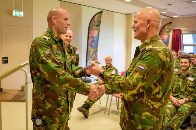 Zwei Männer in militärischer Uniform schütteln sich vor einer sitzenden Gruppe die Hände, mit einem Tisch, Geländer, Bannern, einer Tür, einem Schild, einem Fenster mit Vorhang und Deckenleuchten im Hintergrund.