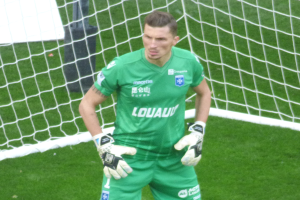 Ein Mann in einem grünen Torwarttrikot steht vor einem Fußballtor, trägt Handschuhe auf einem saftig grünen Feld.