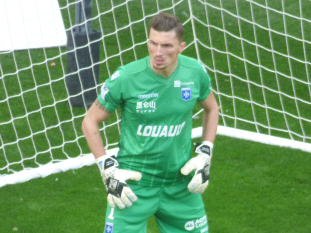 Ein Mann in einem grünen Torwarttrikot steht vor einem Fußballtor, trägt Handschuhe auf einem saftig grünen Feld.