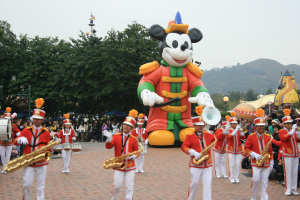 Eine Gruppe von Menschen in Kostümen, die Instrumente spielen, vor einem Mickey-Maus-Floß während eines Umzugs, mit Bäumen, Gebäuden, Hügeln und einem klaren blauen Himmel im Hintergrund und einer Tafel mit Schrift auf der linken Seite.