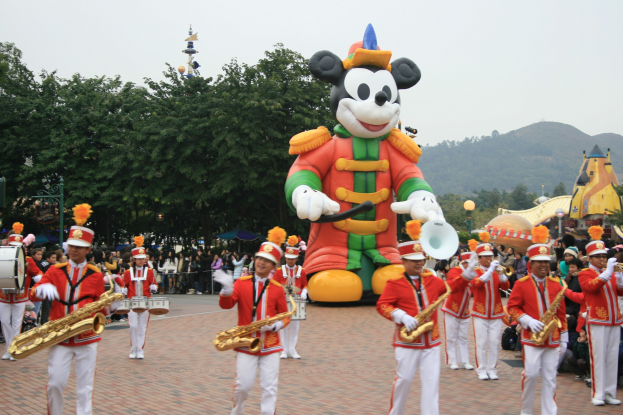 Eine Gruppe von Menschen in Kostümen, die Instrumente spielen, vor einem Mickey-Maus-Floß während eines Umzugs, mit Bäumen, Gebäuden, Hügeln und einem klaren blauen Himmel im Hintergrund und einer Tafel mit Schrift auf der linken Seite.