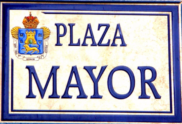 Ein rechteckiges Schild mit weißem Hintergrund, schwarzem Text und blauem Rand, das den Text "Plaza Mayor", ein Wappen-Logo mit einer Krone und fetten Schrift enthält.
