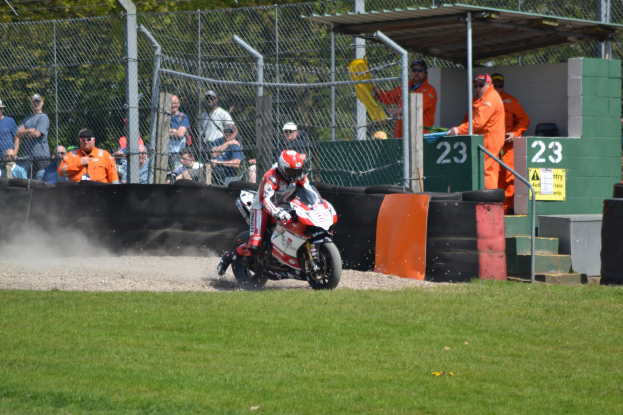 Ein Motorradfahrer in Helm und Handschuhen fährt über ein grasbewachsenes Feld, mit Zuschauern in Mützen und Schutzbrillen, einem eingezäunten Bereich mit Bäumen, Treppen mit Geländern und einem nummerierten Schuppen im Hintergrund.