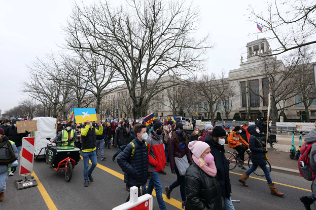 Eine große Gruppe von Menschen nimmt an einer Protestdemo in Washington, D.C. am 21. Januar 2020 teil und marschiert eine Straße entlang, wobei einige Schilder und Banner schwenken und andere Fahrräder fahren, vor einer Kulisse aus Schildern, Bäumen und einem klaren blauen Himmel.