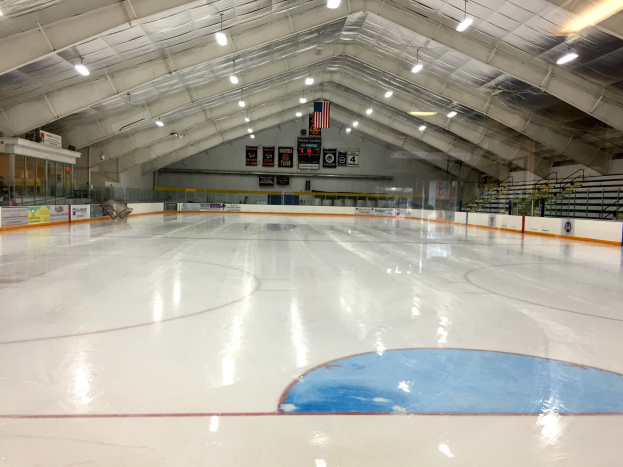 Große Indoor-Eisfläche mit Deckenbeleuchtung, Werbetafeln, einer Rückwand, Treppen mit Geländern und einigen Menschen, was auf eine Sportanlage hinweist, die likely für Hockey genutzt wird.