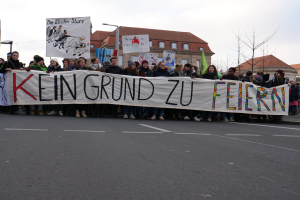Demonstranten mit einem Transparent mit der Aufschrift "Kein Grund zur Feier" gegen deutsche Sparmaßnahmen, mit Straßeninfrastruktur und Gebäuden im Hintergrund bei klarem Himmel.