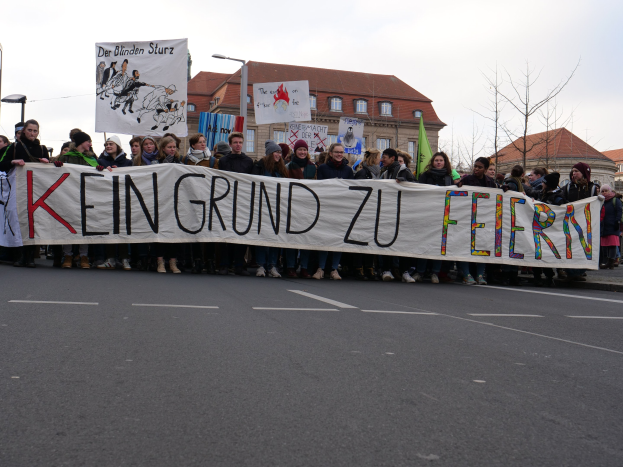Demonstranten mit einem Transparent mit der Aufschrift "Kein Grund zur Feier" gegen deutsche Sparmaßnahmen, mit Straßeninfrastruktur und Gebäuden im Hintergrund bei klarem Himmel.