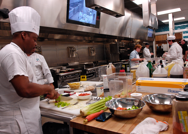 Eine Gruppe von Köchen in einer Küche, die Essen auf einem Schneidbrett zubereiten, mit einem Koch im Vordergrund in einer weißen Schürze und Mütze, der ein Messer hält und von verschiedenen Utensilien, Schüsseln, Gemüse, einem Handy, Flaschen und anderen Gegenständen umgeben ist, während im Hintergrund ein Fernseher und Deckenlampen zu sehen sind.