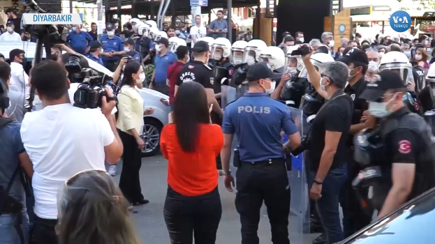 Eine Gruppe von Menschen steht vor einer Reihe von Polizisten in Helmen und mit Kameras, mit Fahrzeugen auf der Straße und Gebäuden, Bäumen und Schildern im Hintergrund, die "türkische Polizei eskaliert Proteste in Ankara" lesen.
