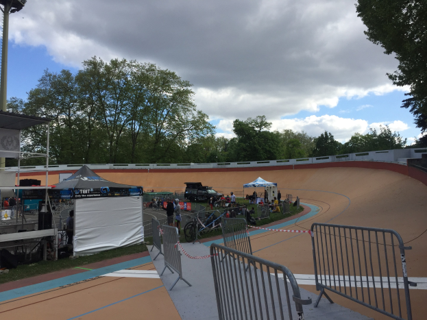 Gruppe von Radfahrern, die auf einem Schotterweg bei einem Outdoor-Event mit Zelten, Fahrzeugen, Geländern, Bäumen und einer bewölkten Himmel fahren.